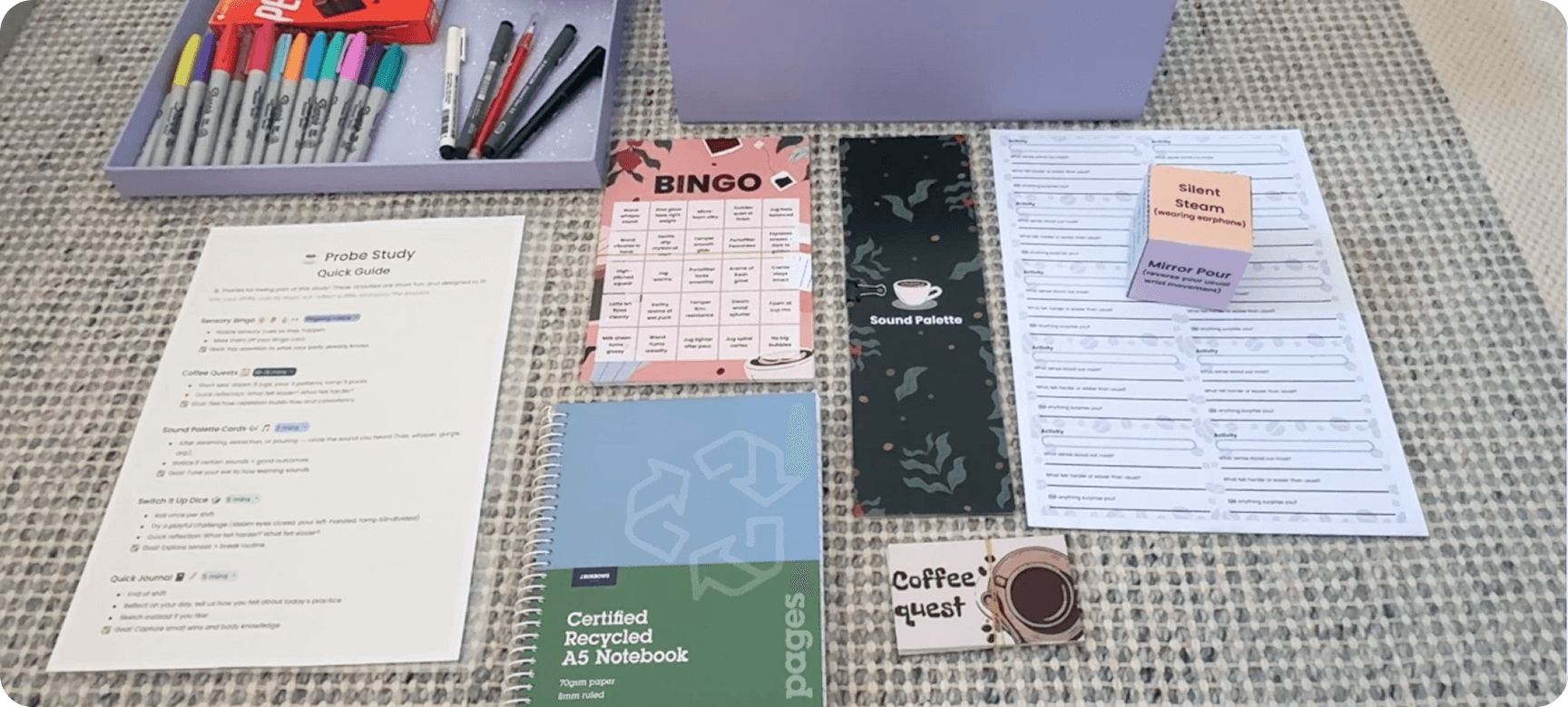 Flat lay of the probe kit in use: a printed quick guide, Sensory Bingo card, Sound Palette cards, Switch-It-Up Dice, Coffee Quest card and a recycled A5 notebook