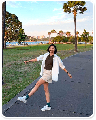 An Ny standing on a path in a Sydney park at sunset, with palm trees and the harbour behind her